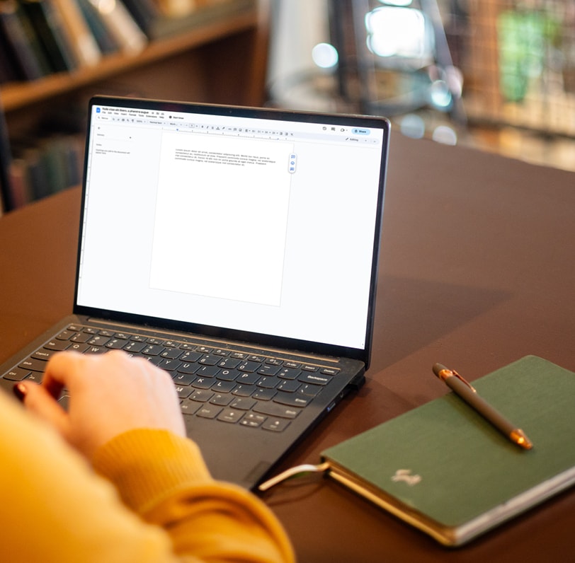 Over-the-shoulder image of a laptop with a journal and pen sitting on the table next to it.