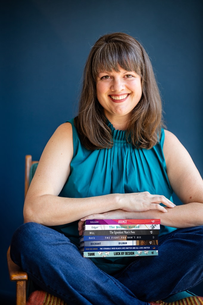 Leadership memoir ghostwriter Amanda Edgar sits cross-legged in a chair holding a stack of books.